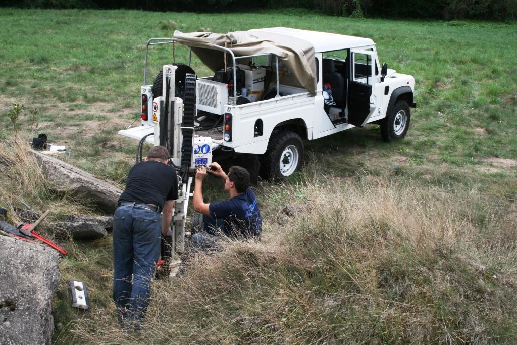 prace terenowe geologów, dwie osoby wykonują sondę za pomoca specjalistycznego urządzenia, na drugim tle stoi terenowy samochów