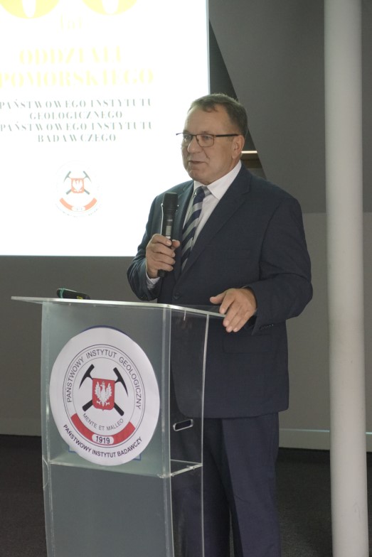 mężczyzna na tle ekranu z prezentacją przemawia do siedzącej w pomieszczeniu publiczności