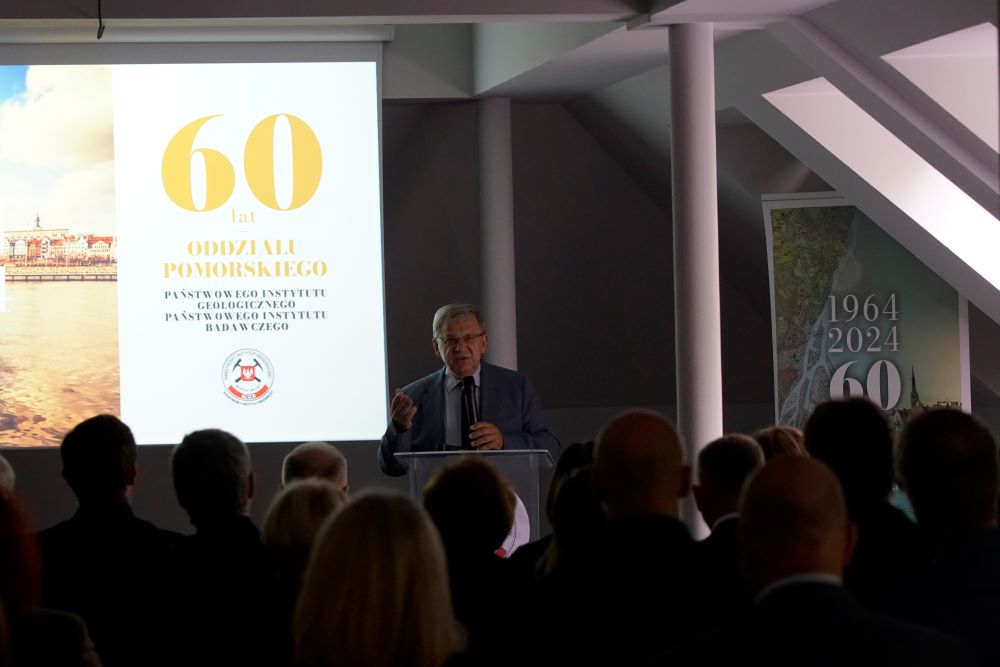 mężczyzna na tle ekranu z prezentacją przemawia do siedzącej w pomieszczeniu publiczności
