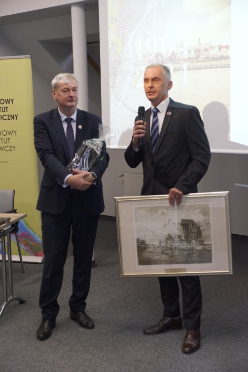 dwóch mężczyzn stoi trzymając w rękach obraz i mikrofon