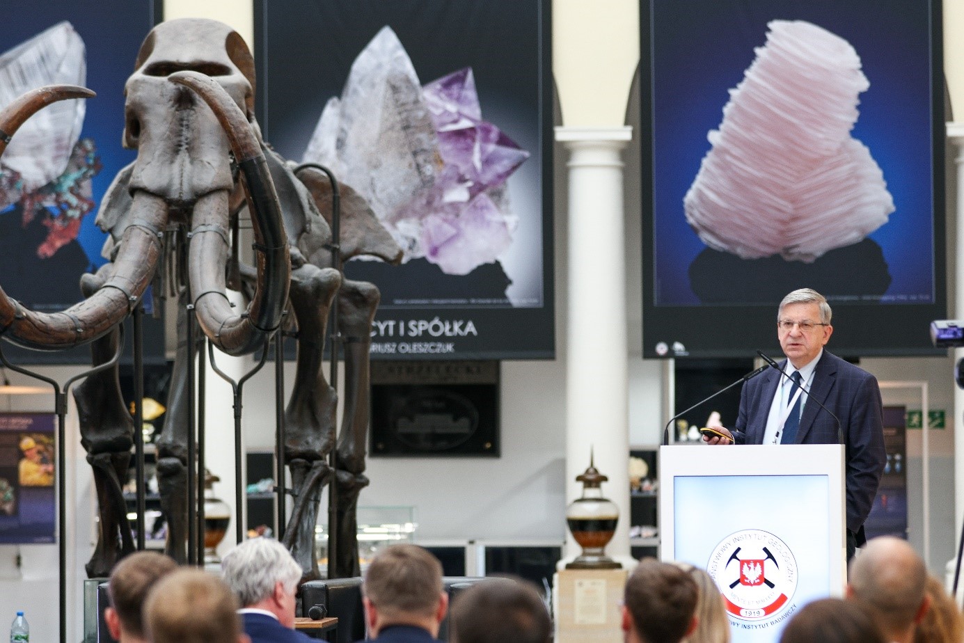 mężczyzna przemawia z mównicy do siedzącej w pomieszczeniu publiczności