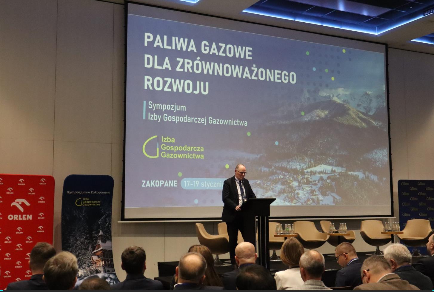 mężczyzna w garniturze przemawia do pełnej sali na tle ekranu z prezentacją
