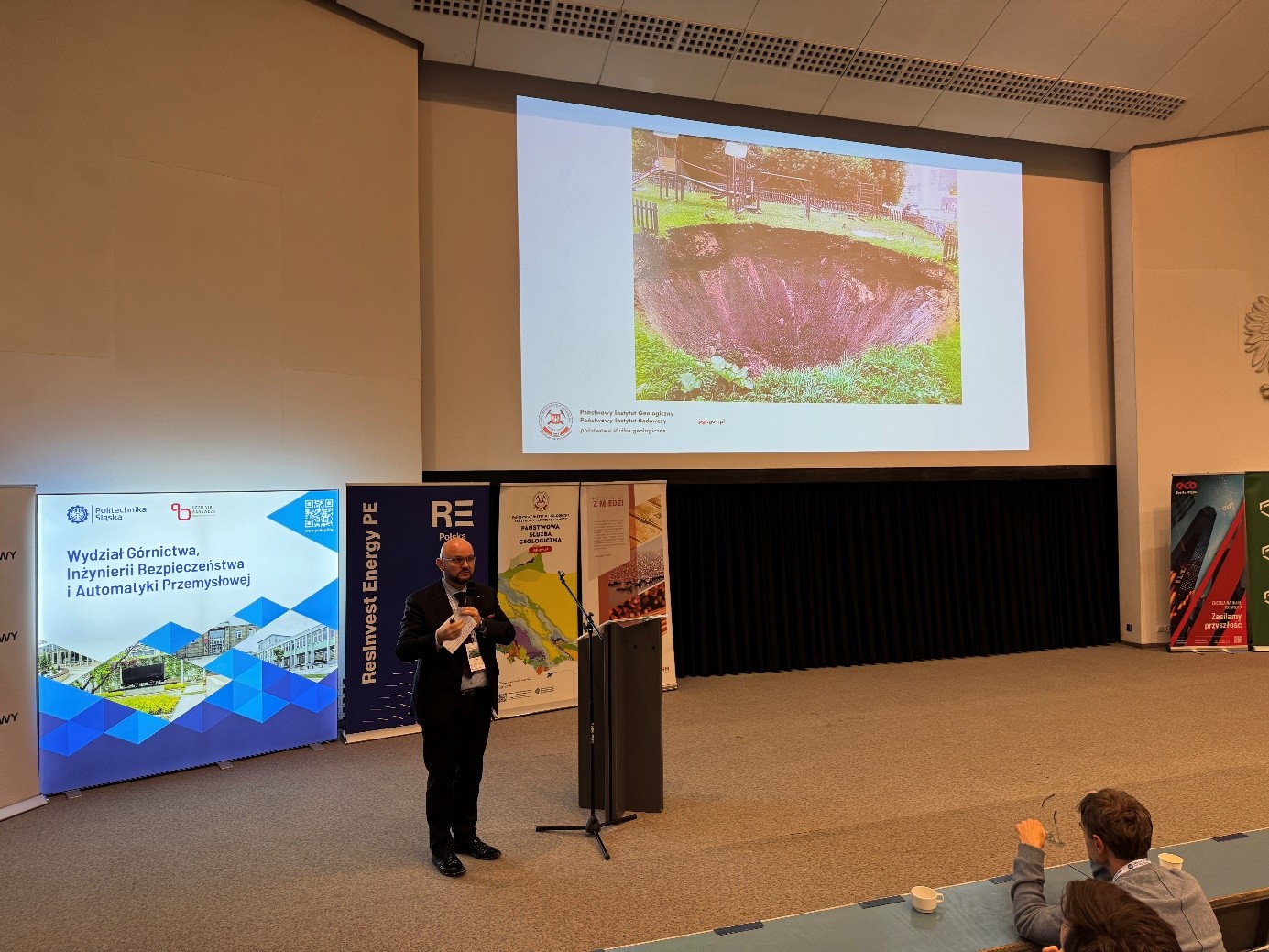 mężczyzna z mikrofonem w ręce podczas prelekcji, na tle ekranu