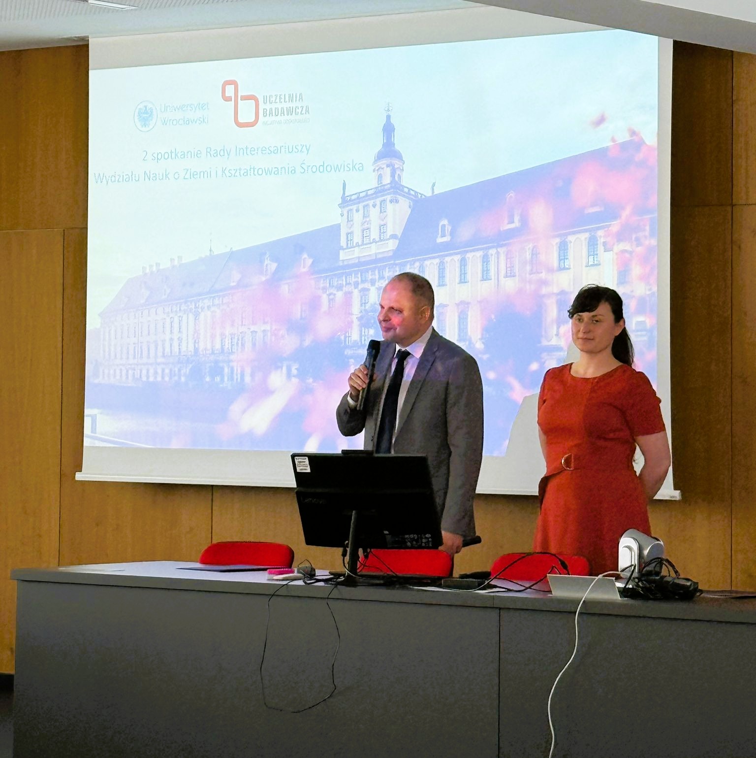 mężczyzna i kobieta stoją za stołem prezydialnym na tle ekranu z prezentacją