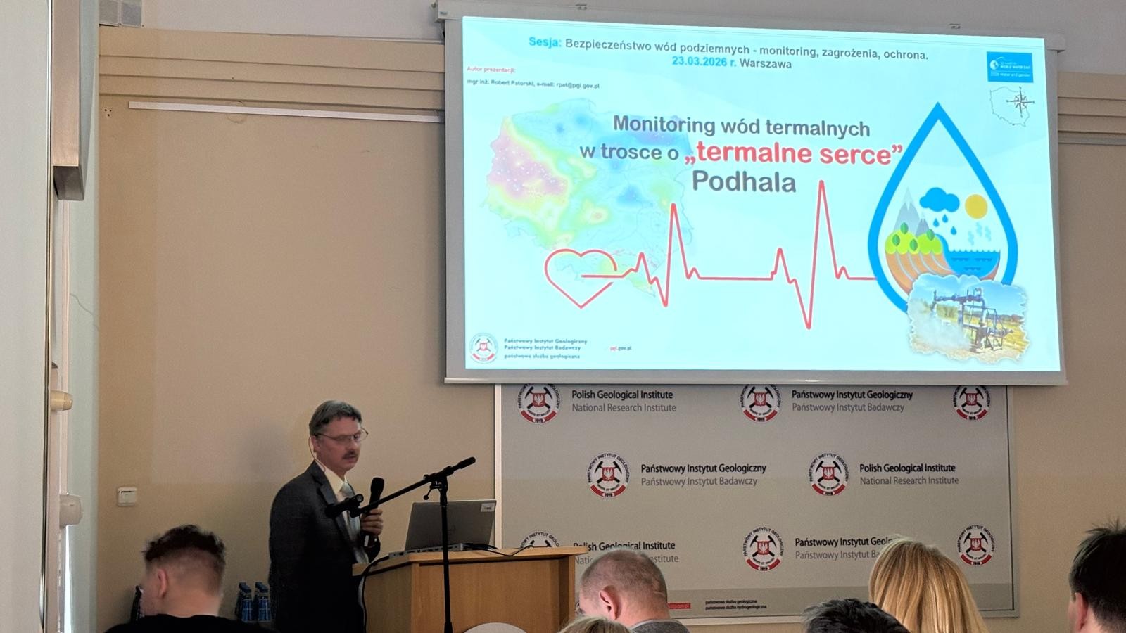 mężczyzna z mikrofonem w ręce podczas prelekcji, na tle ekranu