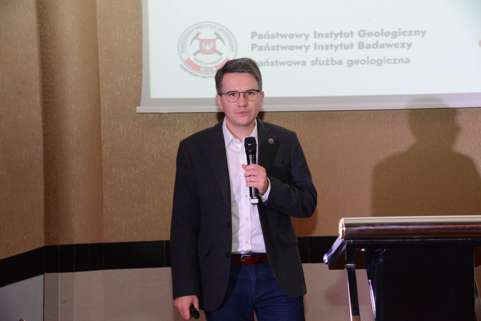 mężczyzna z mikrofonem w ręce podczas prelekcji, na tle ekranu