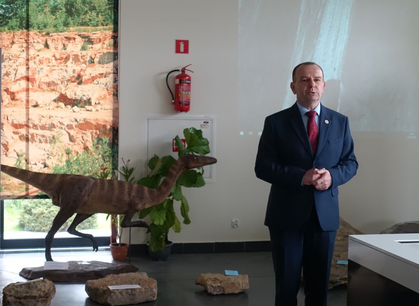 mężczyzna w garniturze przemawia w sali muzeum, na drugim planie makieta dinozaura